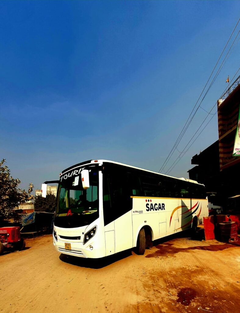 A blue and white bus parked on the side of a mountain road, surrounded by green trees and shrubs. The bus has "Bus On Rent for Chardham Yatra" written in bold letters on its side. The sun is shining on the mountain range in the background, and the sky is blue with a few scattered clouds.