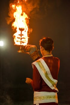 A man in traditional attire holding a flaming arati lamp during a night festival performance.